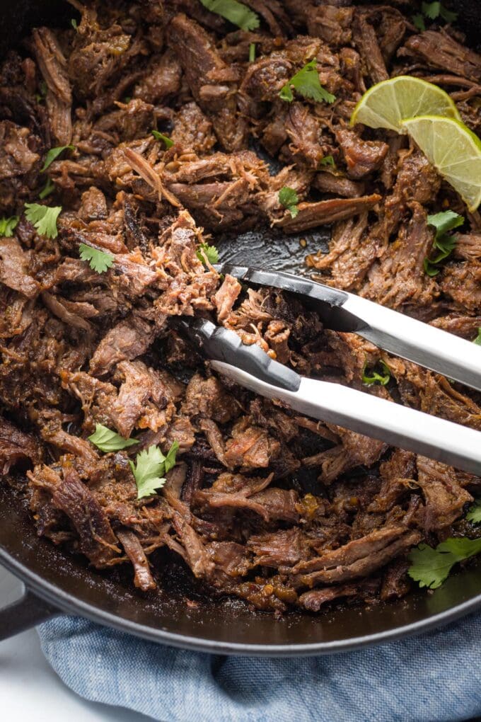 Close-up of tongs serving barbacoa out of a skillet.