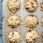 Six blueberry muffins with almond streusel topping in a baking tin.