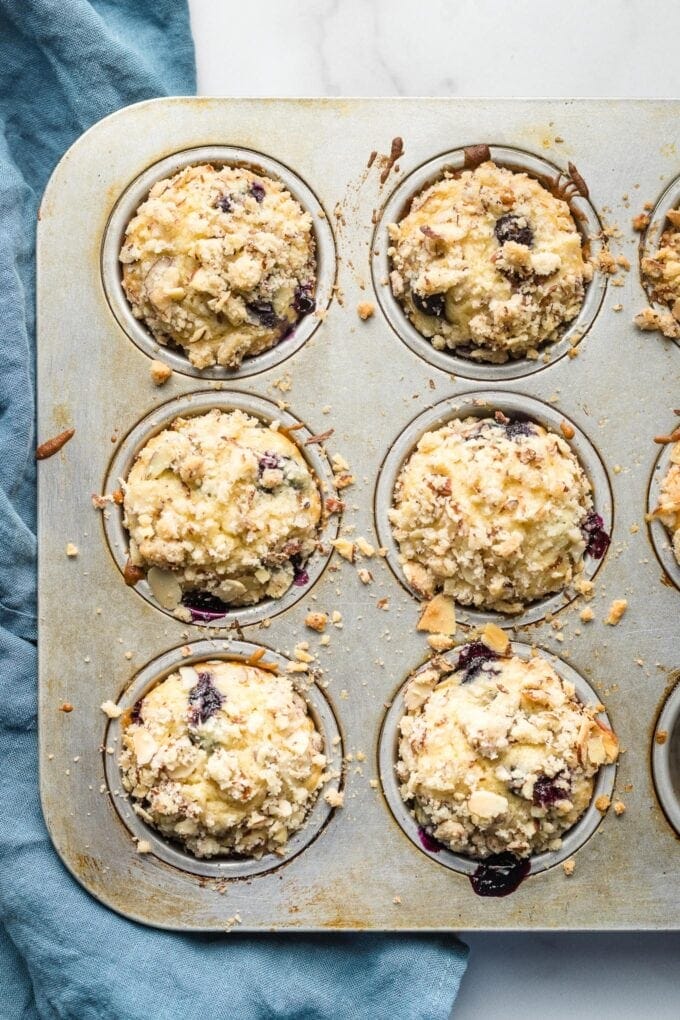 Six blueberry muffins with almond streusel topping in a baking tin.