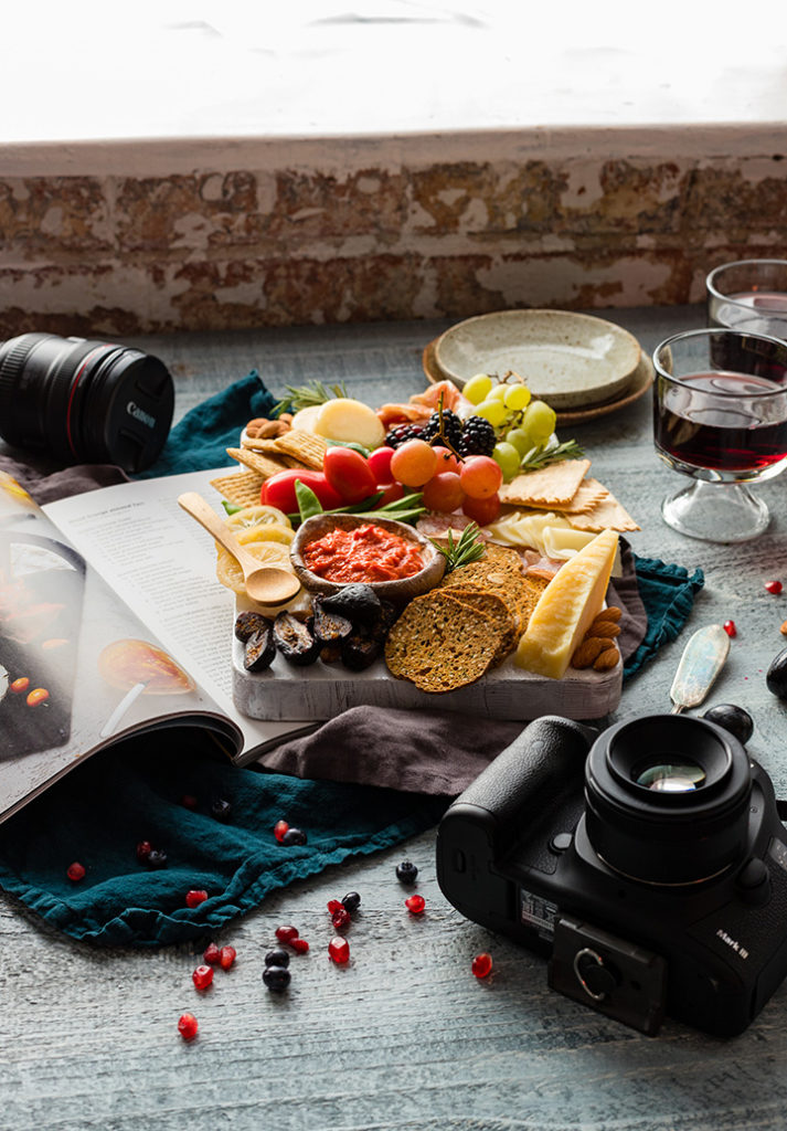 A photography set with a cheese board, open cookbook, glasses of wine, small plates, scattered almonds and other small bites, and Canon camera and lenses.