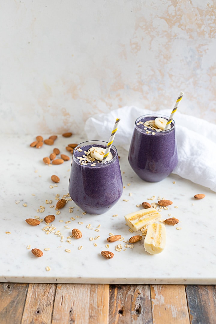 Two banana blueberry almond milk smoothies with banana slices and almonds scattered around.