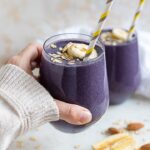 A hand holding a blueberry almond milk smoothie in a small glass with a straw.