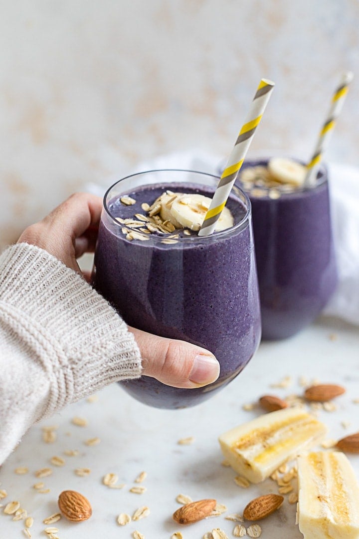A hand holding a blueberry almond milk smoothie in a small glass with a straw.