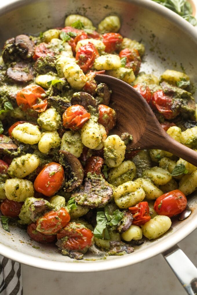 Wooden spoon stirring a large skillet full of pesto gnocchi.