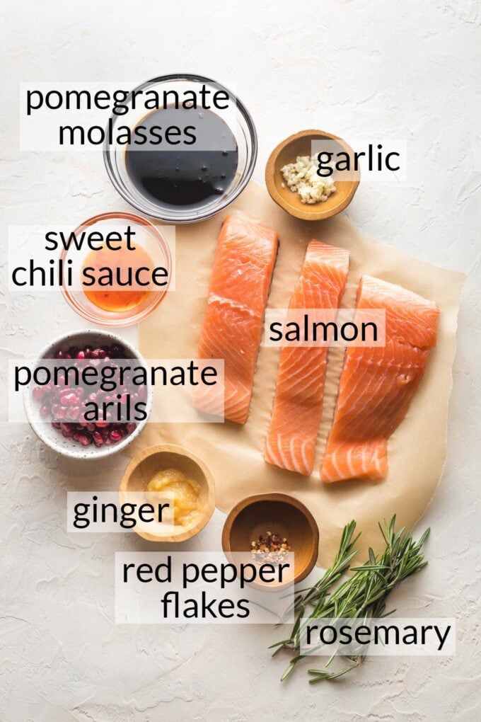 Salmon filets on parchment paper surrounded by prep bowls holding pomegranate molasses, pomegranate arils, sweet chili sauce, garlic, ginger, red pepper flakes, and fresh rosemary.