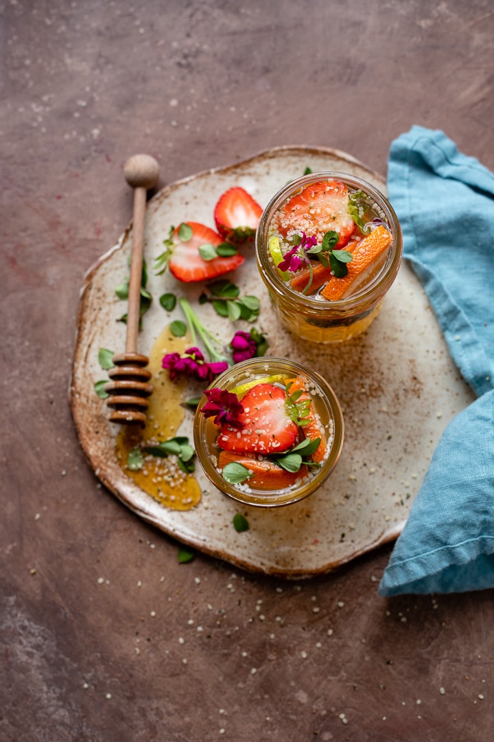 Two tequila kombucha cocktails with fresh herbs and fruit arranged on a small platter with a drizzle of honey.