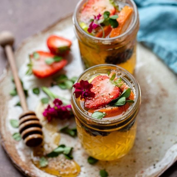 Two tequila kombucha cocktails with fresh herbs and fruit arranged on a small platter with a drizzle of honey.