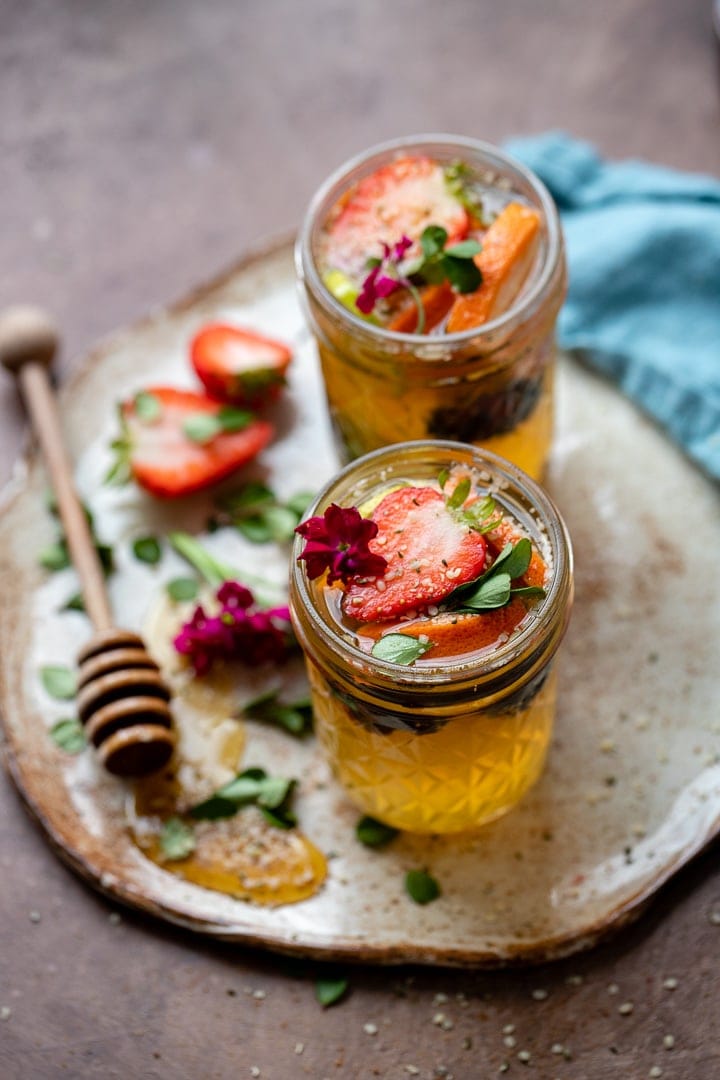 Two tequila kombucha cocktails with fresh herbs and fruit arranged on a small platter with a drizzle of honey.