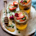 Two tequila kombucha cocktails with fresh herbs and fruit arranged on a small platter with a drizzle of honey.