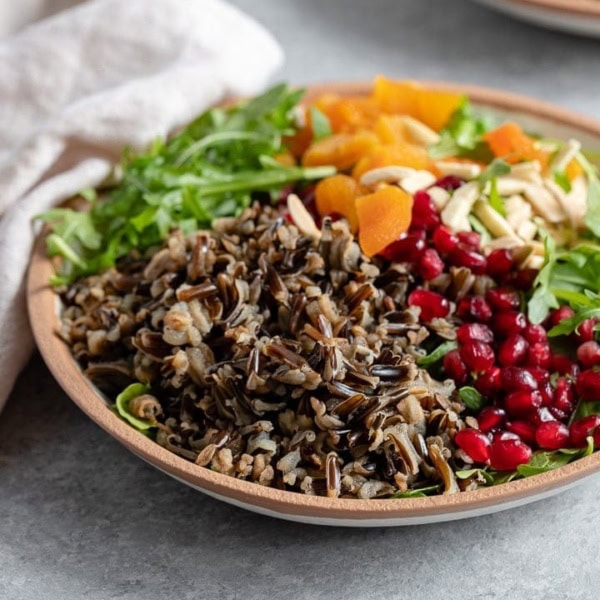 A bowl of wild rice winter salad with arugula, pomegranate arils, almonds, and dried apricots.