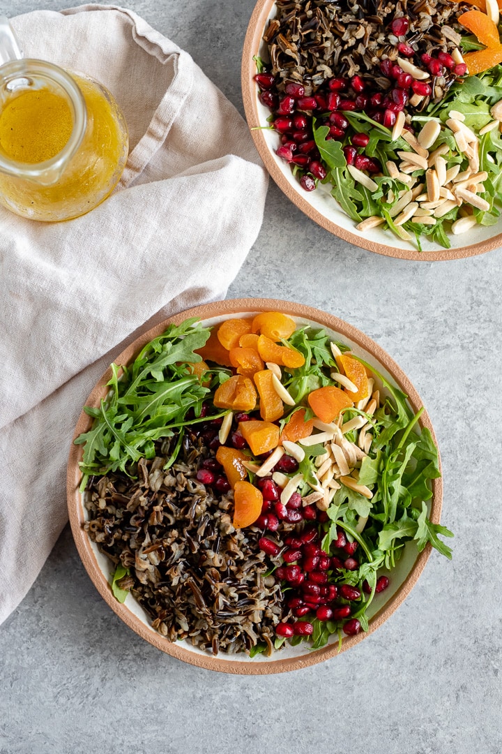 A bowl of wild rice winter salad with arugula, pomegranate arils, almonds, and dried apricots.