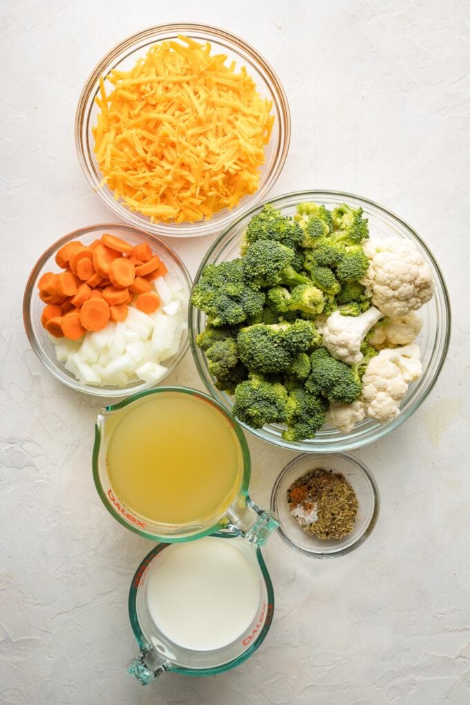 Prep bowls holding shredded cheddar cheese, carrots, onions, broccoli, cauliflower, broth, milk, and seasonings.