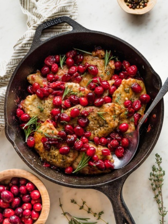 Cast iron skillet containing a balsamic cranberry chicken dish garnished with fresh thyme and rosemary.