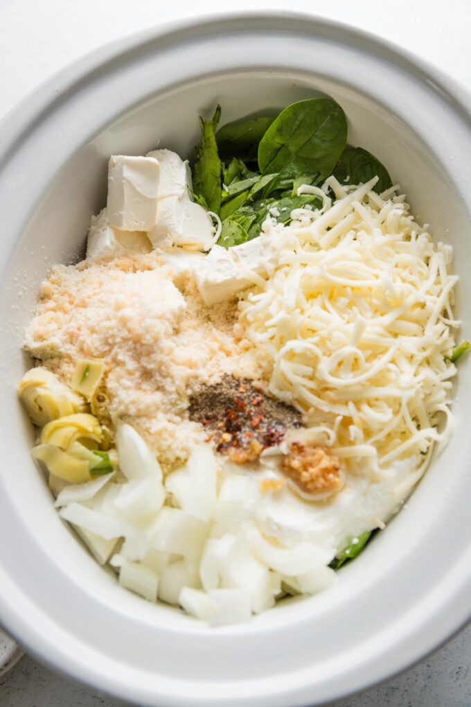 Small Crockpot bowl filled with fresh baby spinach, cream cheese, Greek yogurt, mozzarella, Parmesan, onion, garlic, and spices.