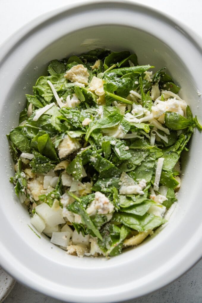 Spinach, cream cheese, artichokes, and other ingredients all stirred together in the bowl of a Crockpot.