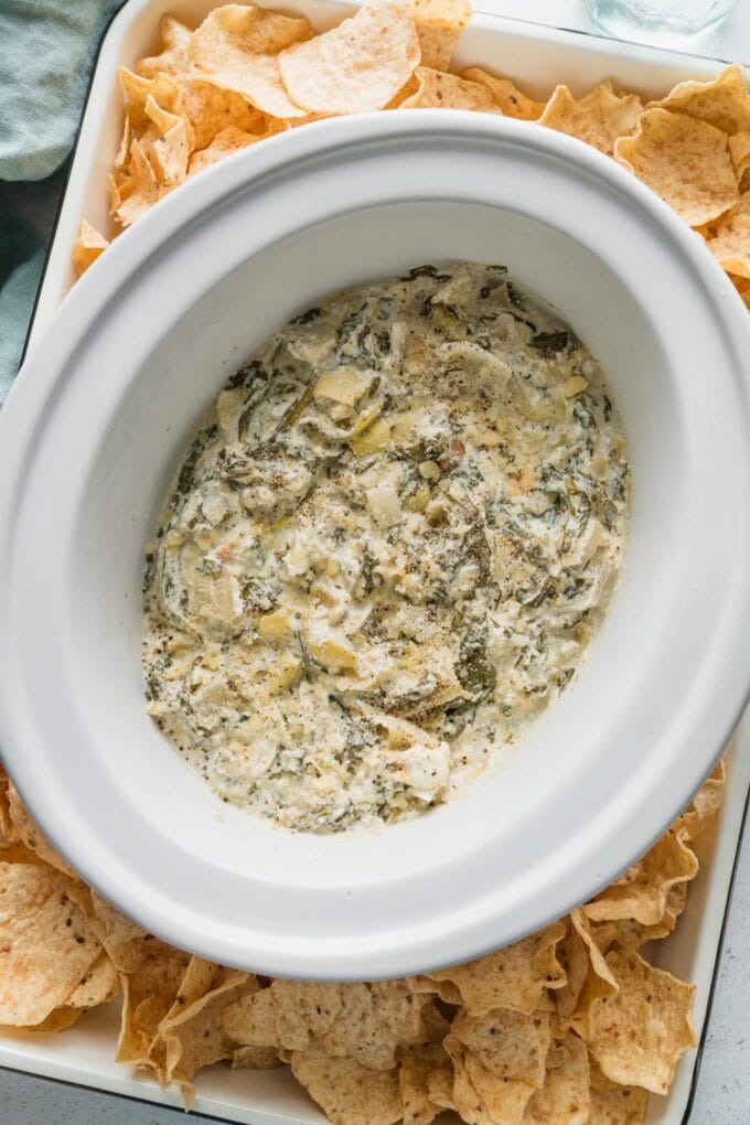 Small Crockpot full of spinach artichoke dip on a tray of tortilla scoops.