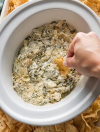 Woman's hand scooping a tortilla chip into a crockpot full of spinach artichoke dip.