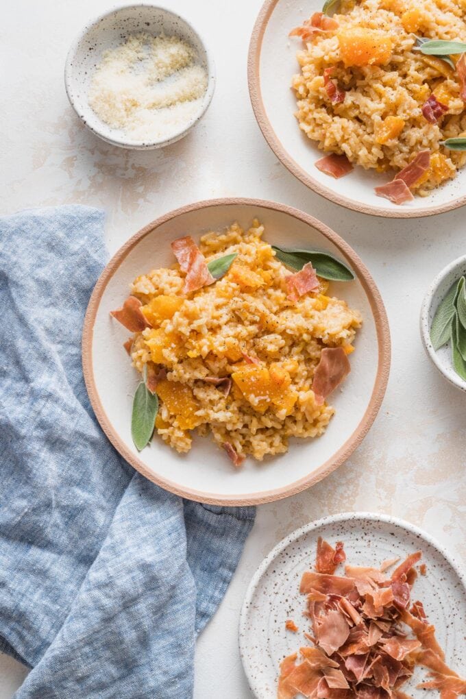 Bowls of Instant Pot butternut squash risotto served with crisped prosciutto and generous sprinkles of Parmesan cheese.