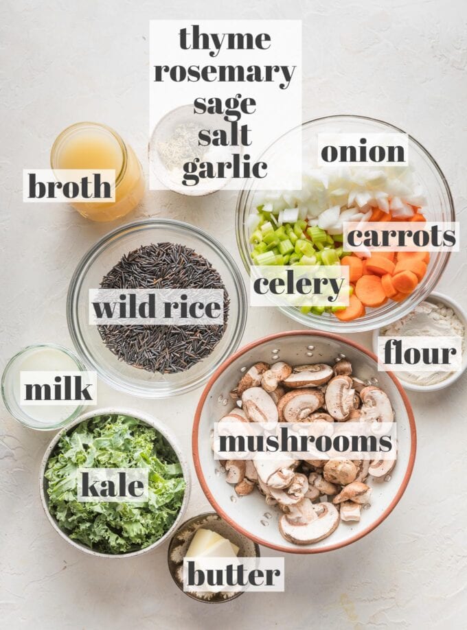 Labeled overhead photo of wild rice, sliced mushrooms, carrots, onion, celery, minced garlic, dried sage, thyme, and rosemary, salt, broth, milk, butter, flour, and kale arranged in prep bowls and ready to cook.