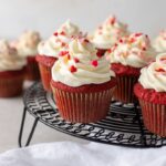 One-bowl red velvet cupcakes, baked and frosted with heart sprinkles, arranged on a black cooling rack.