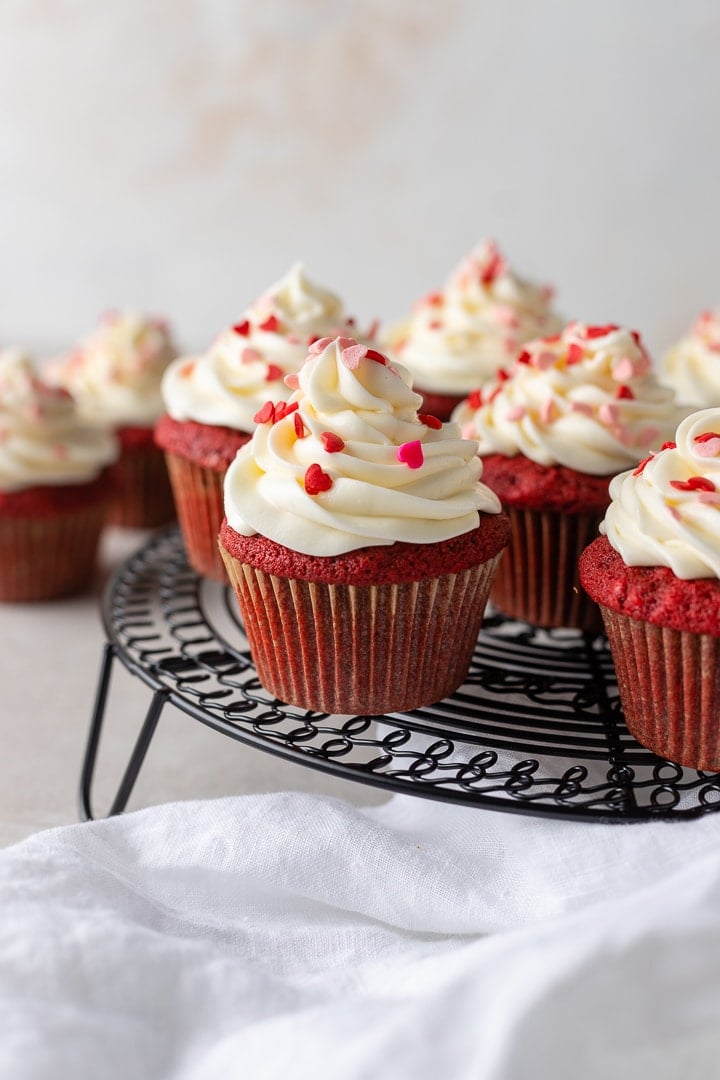 One-bowl red velvet cupcakes, baked and frosted with heart sprinkles, arranged on a black cooling rack.