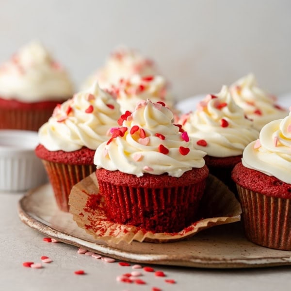 Close-up of an unwrapped red velvet cupcake with cream cheese frosting and heart sprinkles.