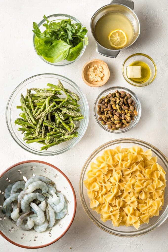 Prep bowls containing shrimp, pasta, asparagus, pistachios, herbs, lemon juice, butter, and garlic.