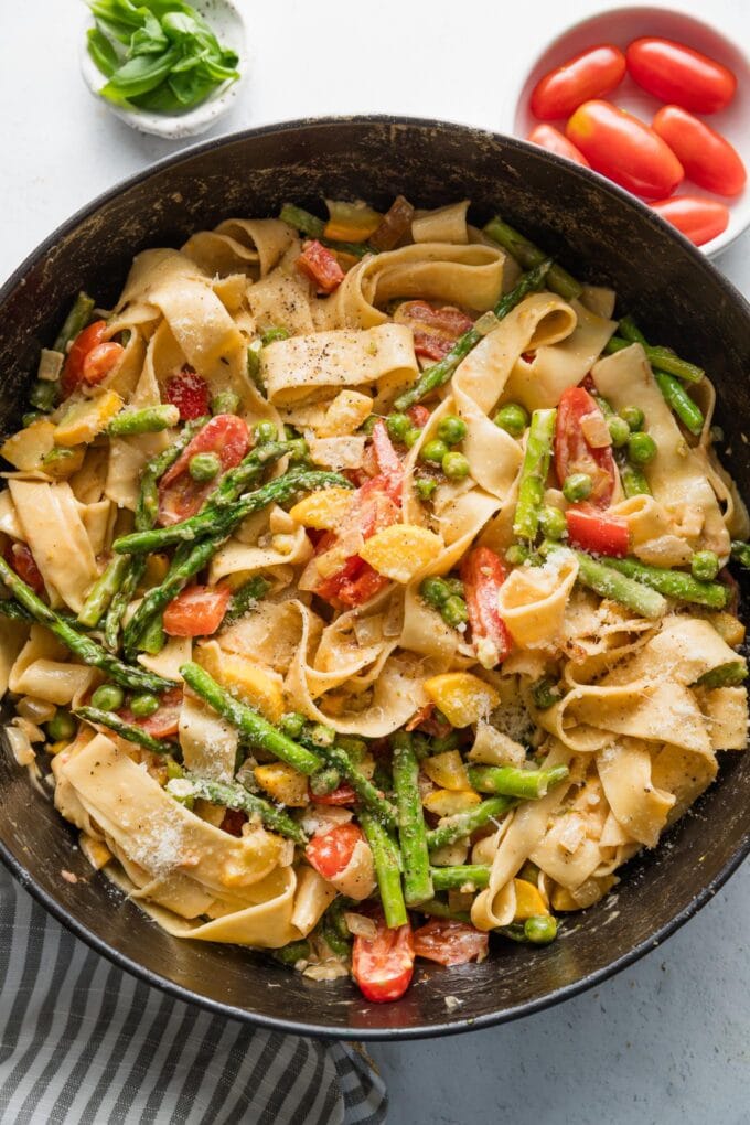 Large Staub cast iron skillet filled with a homemade pasta primavera in a light cream sauce, with Parmesan, basil, and cherry tomatoes scattered on a kitchen counter nearby.