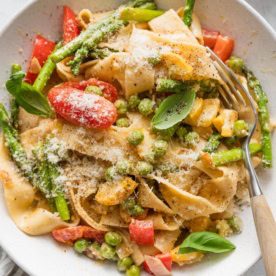 Close up overhead image of a low pasta bowl filled with a creamy pasta primavera, made with ribbons of pappardelle, asparagus, tomatoes, peppers, peas, and yellow squash in a light homemade cream sauce.