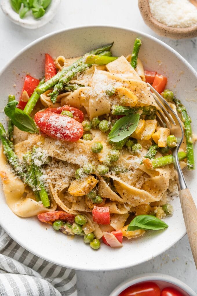 Close up overhead image of a low pasta bowl filled with a creamy pasta primavera, made with ribbons of pappardelle, asparagus, tomatoes, peppers, peas, and yellow squash in a light homemade cream sauce.