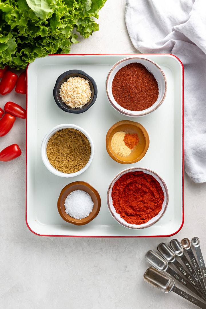 Small bowls sitting on a white tray, each containing one or two of the spices used in homemade taco seasoning: chili powder, paprika, cumin, dried minced onion, kosher salt, garlic powder, cayenne pepper.