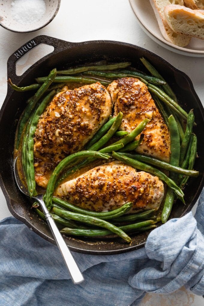 Overhead view of a serving utensil about to lift out a portion of honey mustard chicken and green beans from a black cast iron skillet.