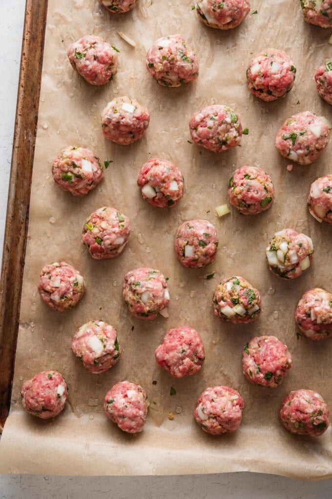 Rimmed baking sheet lined with parchment paper and filled with uncooked lamb meatballs.