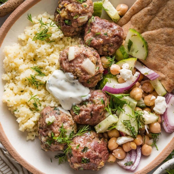 Bowl of Moroccan-inspired lamb meatballs served with couscous, a cucumber chickpea salad, tzatziki sauce, pita bread, and fresh dill.