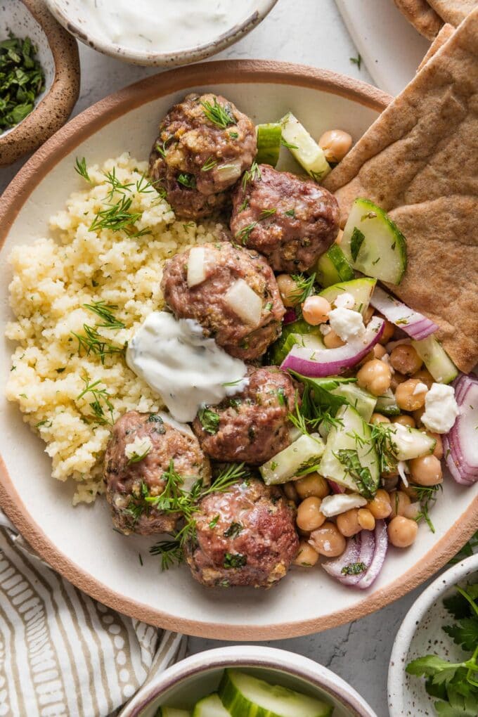 Bowl of Moroccan-inspired lamb meatballs served with couscous, a cucumber chickpea salad, tzatziki sauce, pita bread, and fresh dill.
