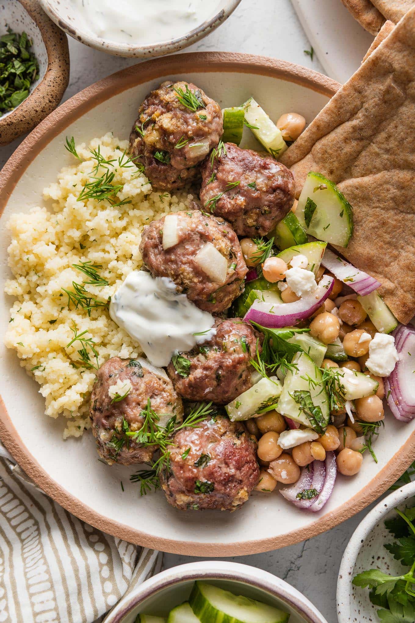 Bowl of Moroccan-inspired lamb meatballs served with couscous, a cucumber chickpea salad, tzatziki sauce, pita bread, and fresh dill.