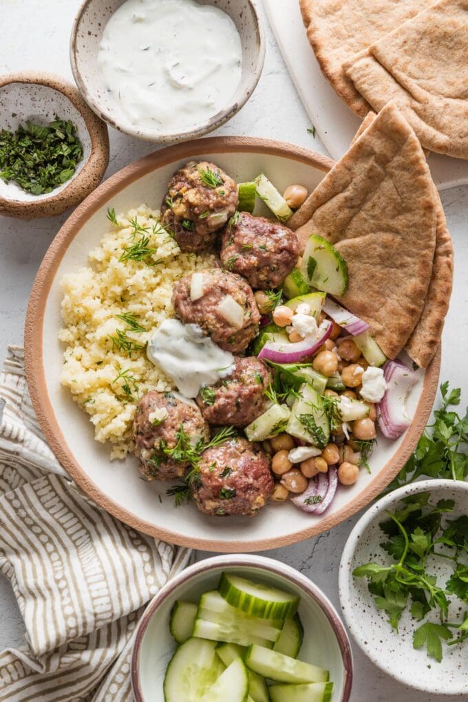 Bowl full of lamb meatballs seasoned with cinnamon, cumin, and a hint of cayenne pepper, served with pita, couscous, and a chickpea cucumber salad, with extra salad fixings, herbs, and sauce nearby.