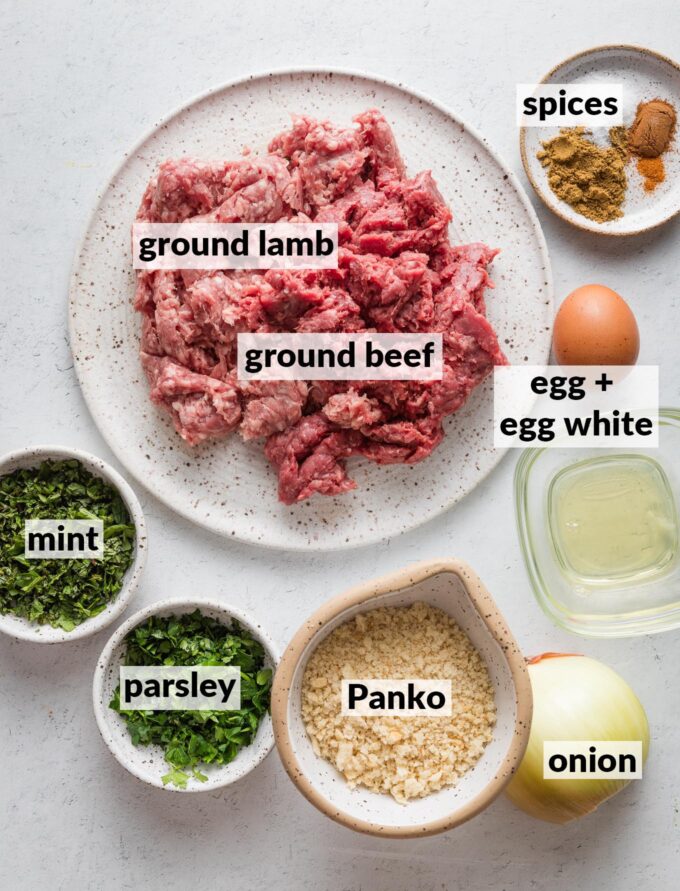 Labeled overhead image of ground lamb, ground beef, spices, one whole egg plus one egg white, Panko, yellow onion, fresh mint and fresh parsley.