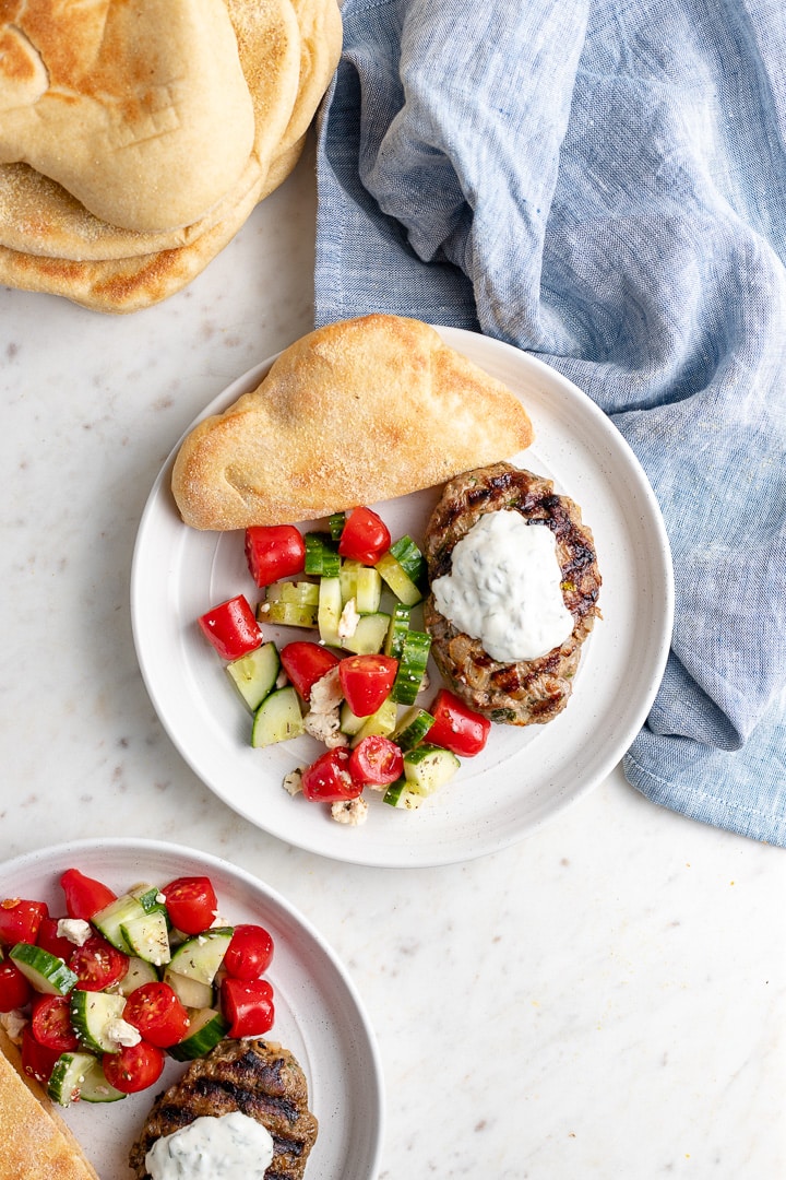 A plate with a Moroccan lamb burger, yogurt sauce, tomato cucumber salad, and fresh pita bread.