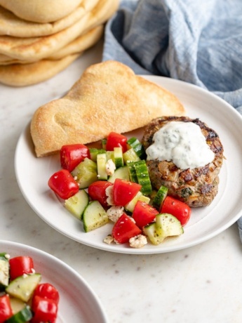 A plate with a Moroccan lamb burger, yogurt sauce, tomato cucumber salad, and fresh pita bread.