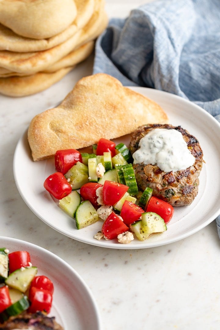 A plate with a Moroccan lamb burger, yogurt sauce, tomato cucumber salad, and fresh pita bread.