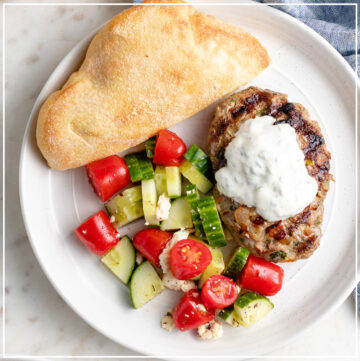 A plate with a Moroccan lamb burger, yogurt sauce, tomato cucumber salad, and fresh pita bread.