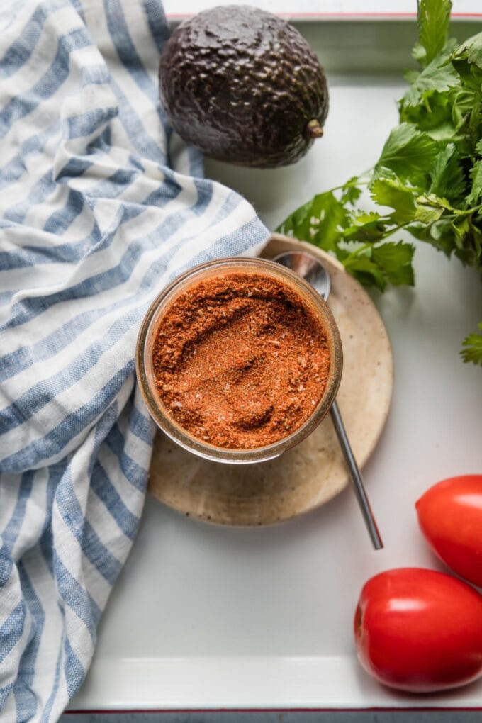 Jar of homemade taco seasoning with a small spoon nearby, tomatoes, and green leaf lettuce.