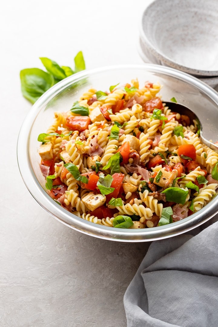 Large clear Pyrex bowl filled with a Caprese Pasta Salad including rotini, tomatoes, mozzarella, basil, prosciutto, pine nuts, and dressing.
