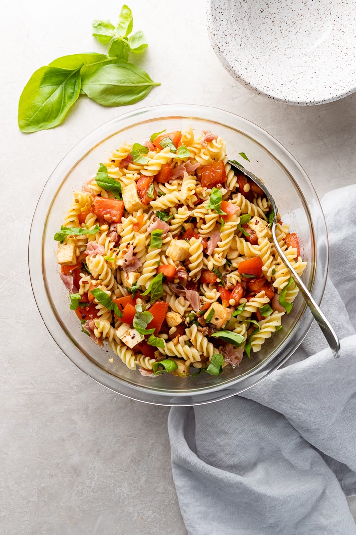 Large clear Pyrex bowl filled with a Caprese Pasta Salad including rotini, tomatoes, mozzarella, basil, prosciutto, pine nuts, and dressing.