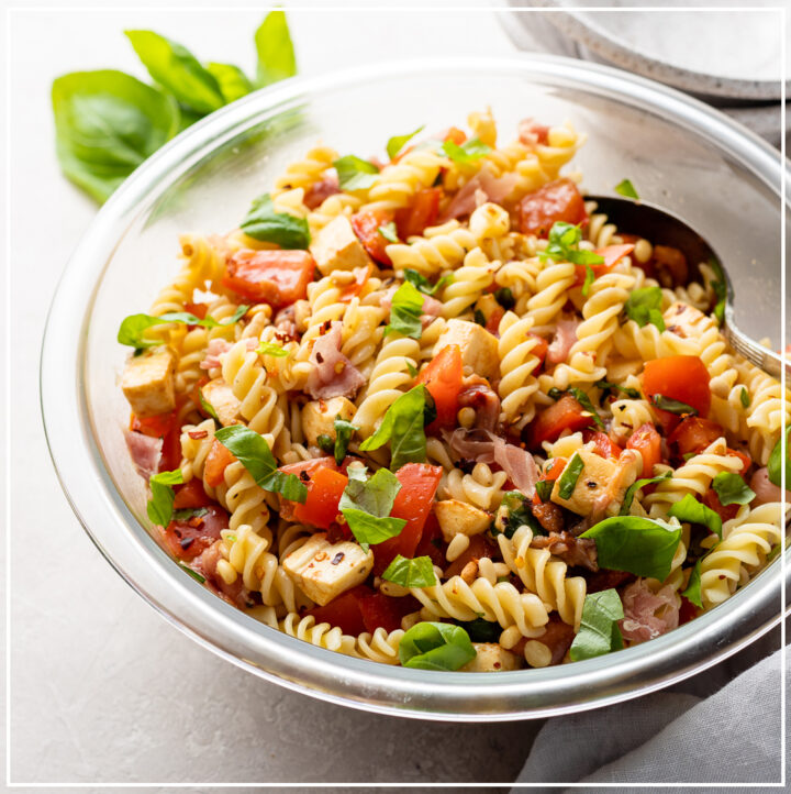 Large clear Pyrex bowl filled with a Caprese Pasta Salad including rotini, tomatoes, mozzarella, basil, prosciutto, pine nuts, and dressing.