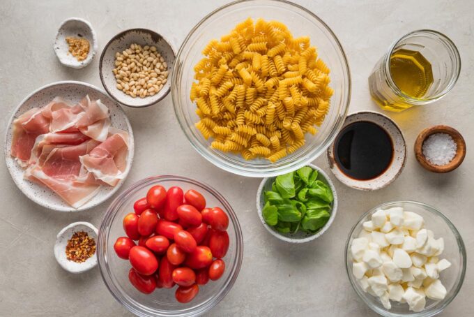 Flat lay image of all the ingredients for an elevated caprese pasta salad with prosciutto, fresh basil, and pine nuts.
