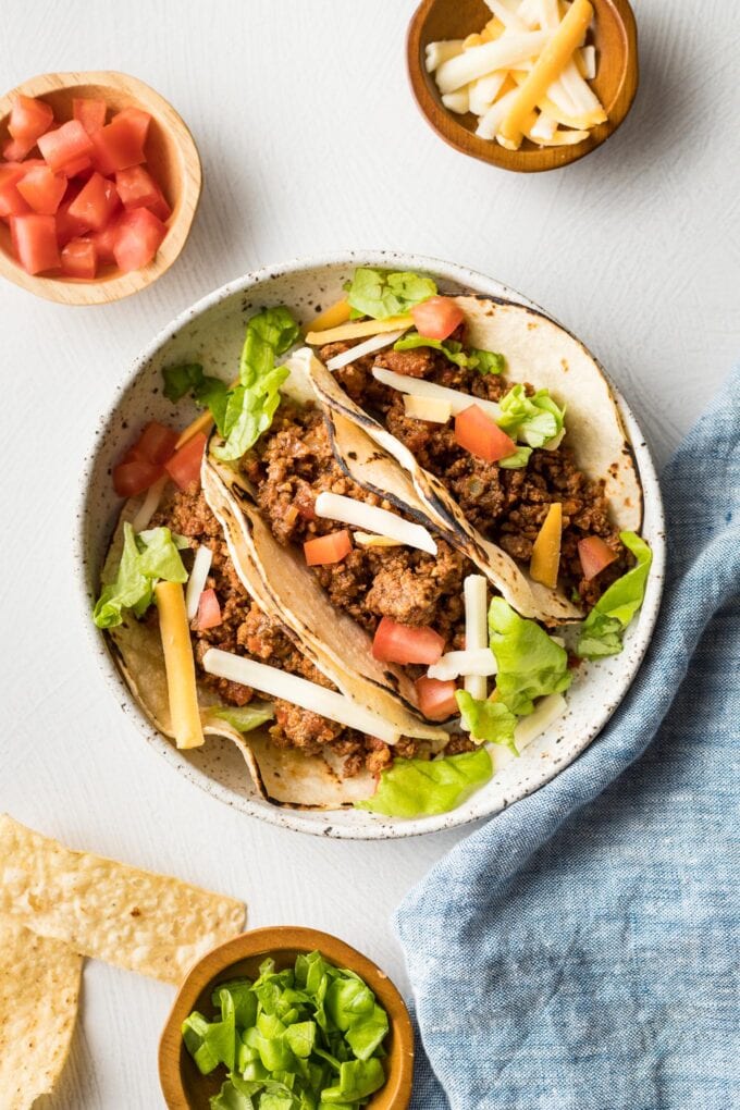 Plate with ground beef tacos and small bowls of tomatoes, cheese, and lettuce.