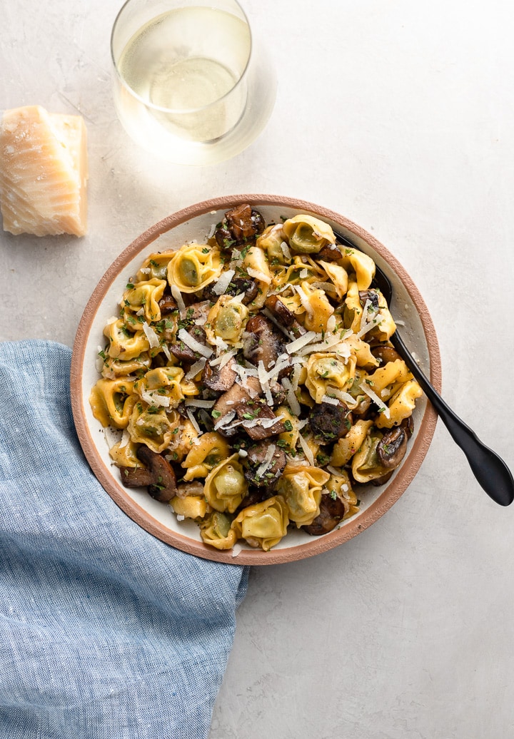 A small pasta bowl filled with tortellini with mushrooms and Parmesan cheese, with more Parmesan and a glass of white wine to the side.