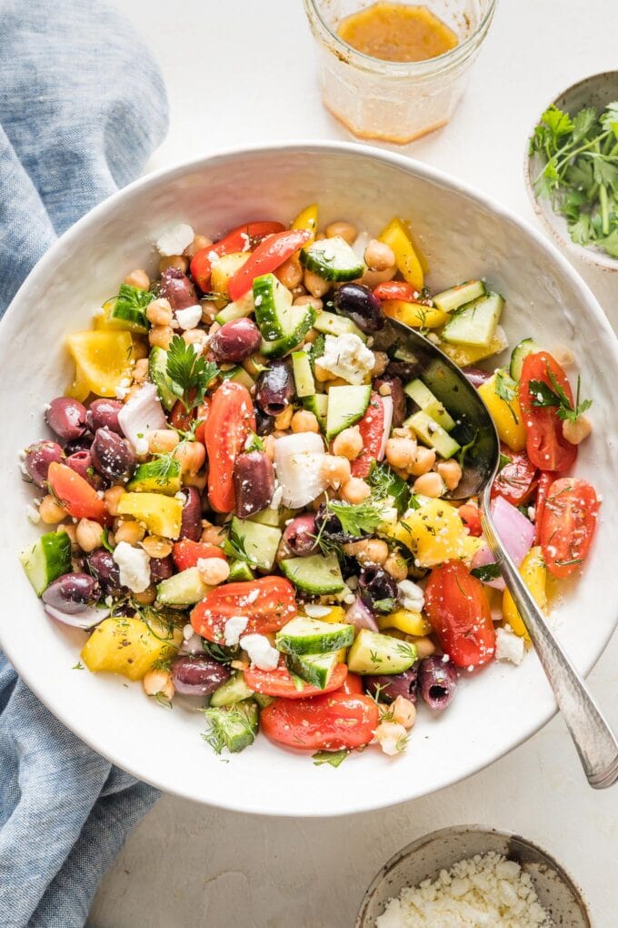 Serving spoon stirring a Greek chickpea salad in a large white ceramic bowl.